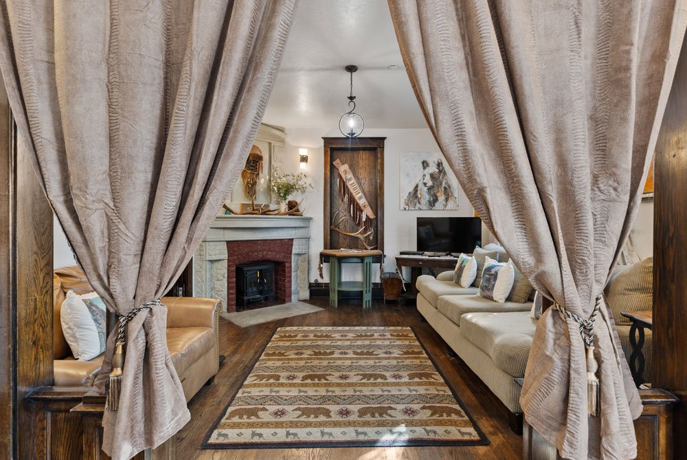 A cozy living room with tan curtains, a patterned area rug with bears, a beige sofa, a leather armchair, a brick fireplace, a TV, and rustic decor on wood floors.