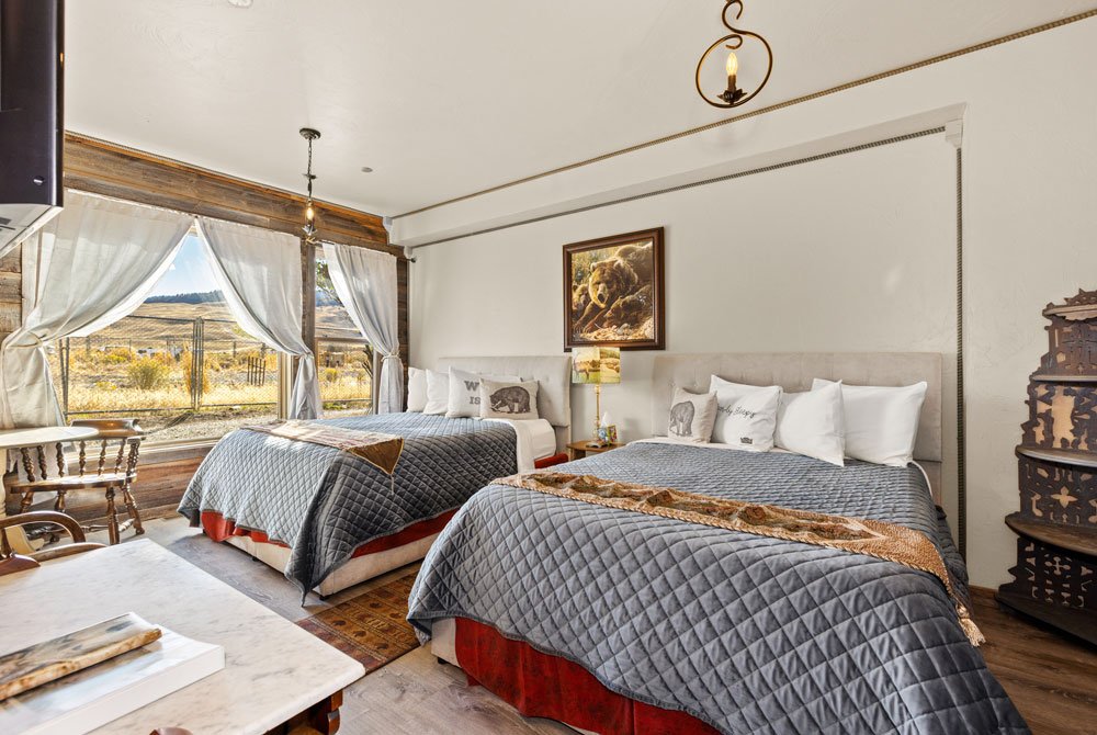 Rustic bedroom with two double beds, gray quilted bedding, animal-themed pillows, wood furniture, and large windows with sheer curtains showing a scenic outdoor view. Warm, cozy decor and natural light fill the space.