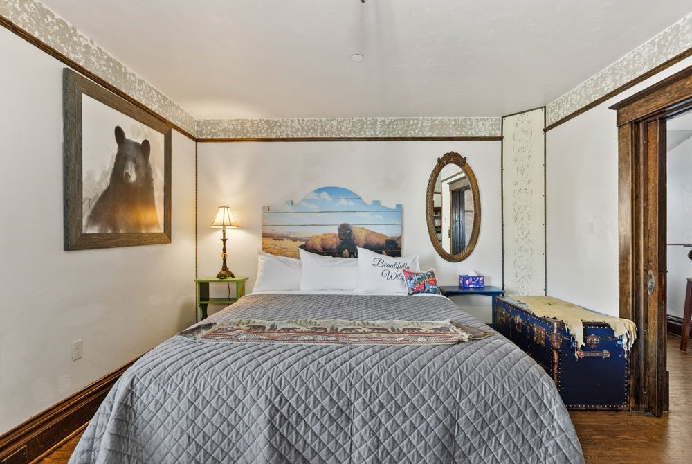 A cozy bedroom with a neatly made bed, a bear photo on the wall, a lamp on a small table, an oval mirror, a mural of rocks and ocean, and a vintage trunk at the foot of the bed.