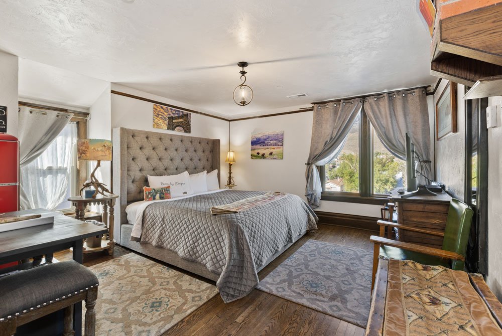 A cozy bedroom with a large bed, tufted headboard, patterned rug, wooden floors, a desk, armchair, two lamps, and large windows with gray curtains letting in natural light.
