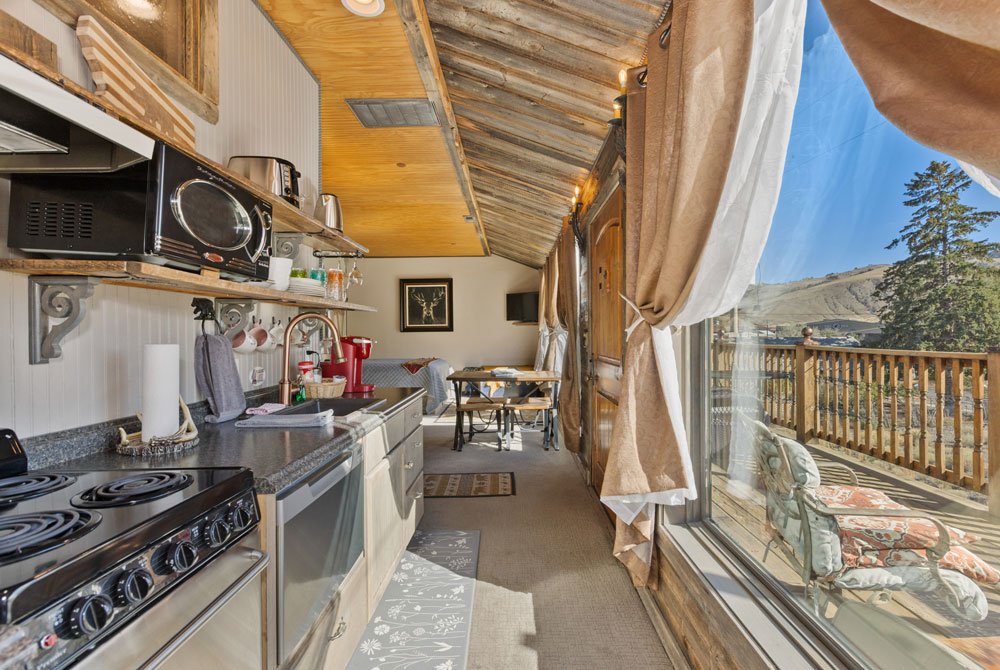 A cozy, narrow kitchen with rustic decor features a stove, microwave, and sink on one side. Large windows and curtains reveal a sunny deck with chairs and a scenic view of hills and trees outside.