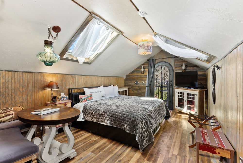 Cozy attic bedroom with sloped ceilings, skylights, a large bed with gray bedding, rustic wood paneling, a round table with chairs, TV on a white cabinet, and decorative lighting fixtures.