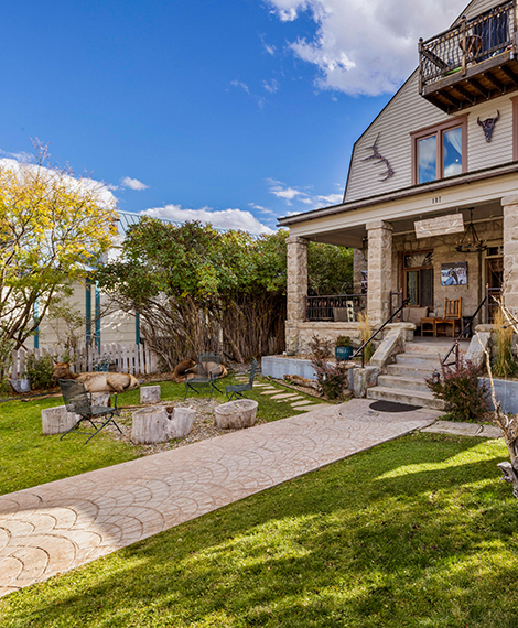 A stone pathway leads to a house with a covered porch, stone columns, and a balcony. The yard has green grass, trees, and outdoor seating. The sky is blue with scattered clouds.