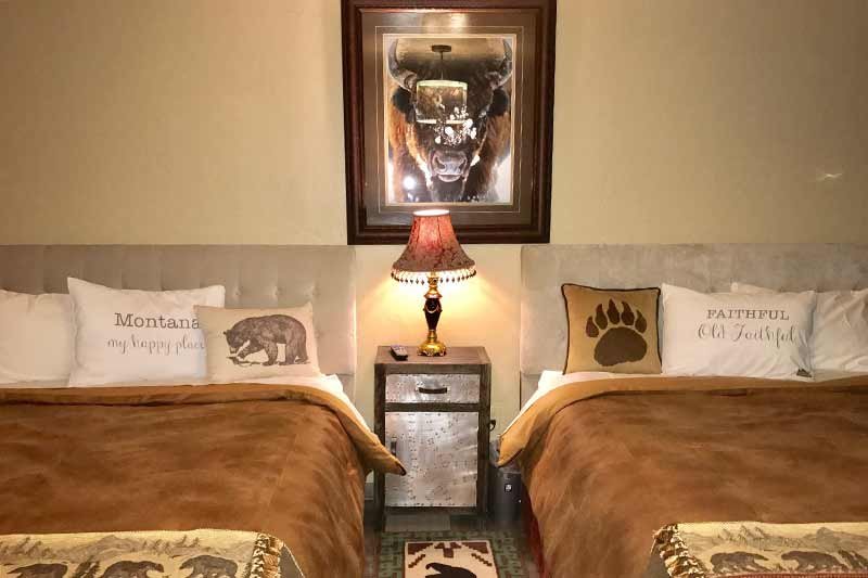 A cozy bedroom with two beds covered in brown blankets, animal-themed pillows, a nightstand with a lamp in between, and a framed buffalo picture on the wall above the lamp.