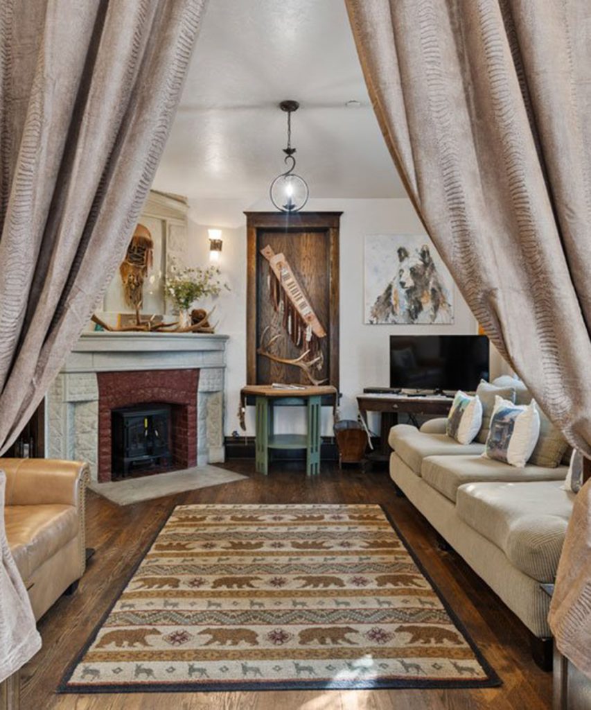 Cozy living room with beige couches, a patterned rug featuring bears, a fireplace, and a TV. Neutral curtains frame the scene, and rustic decor accents the space, including antlers and woodland-themed artwork.