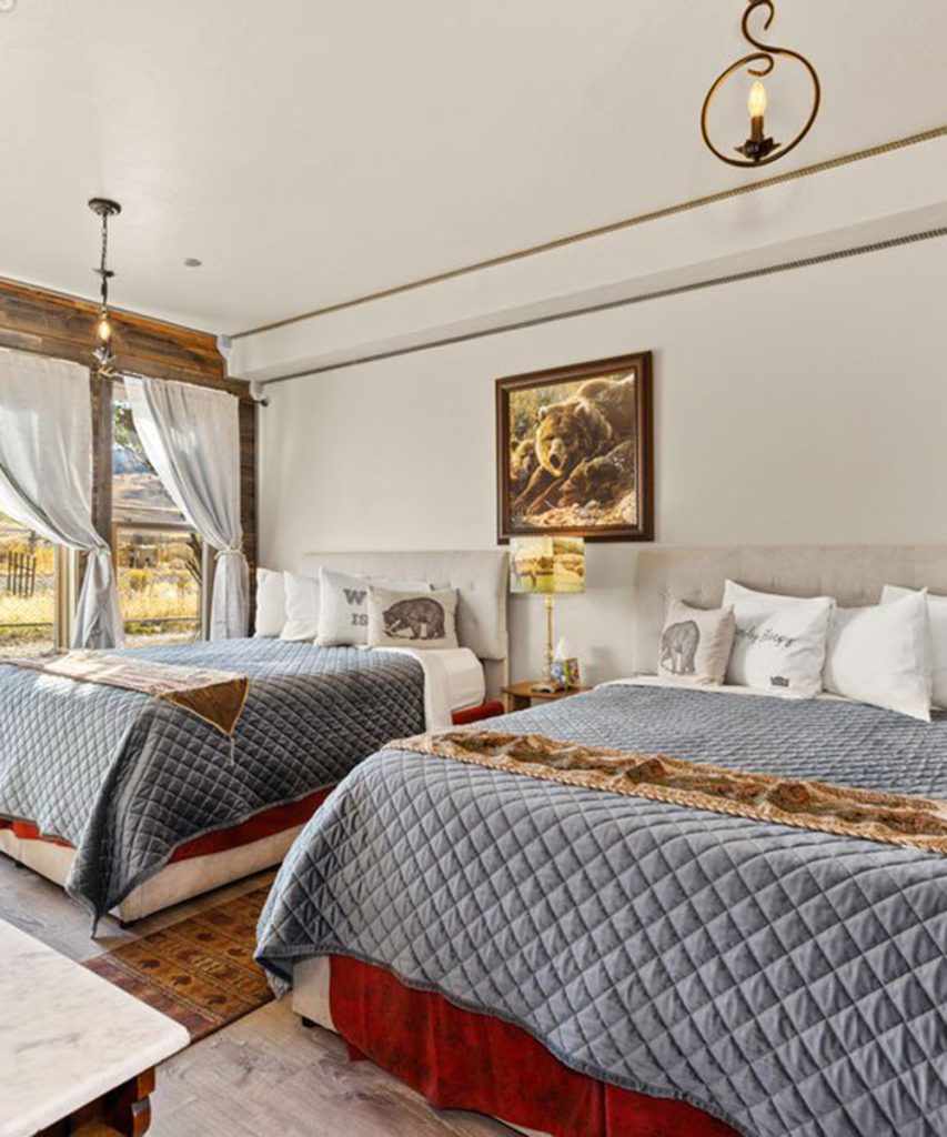 A cozy bedroom with two double beds, blue quilted bedspreads, animal-themed pillows, a bear painting on the wall, a rustic light fixture, and large windows with sheer curtains showing an outdoor view.