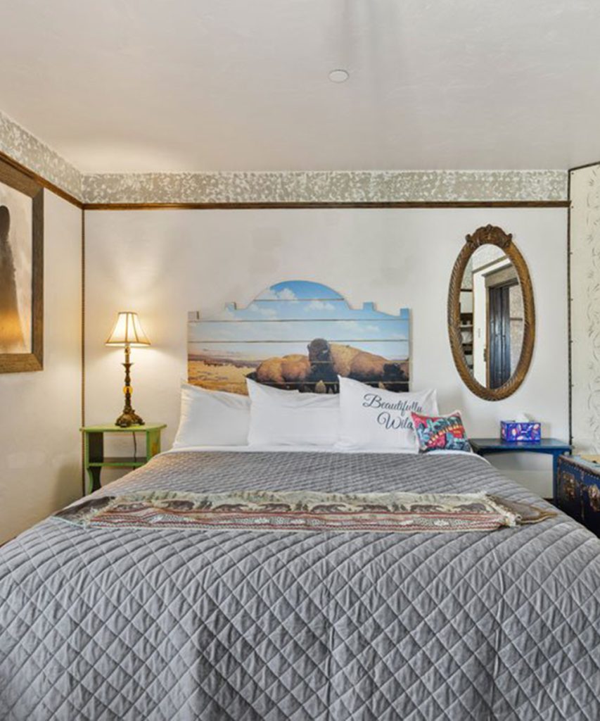 A cozy bedroom with a neatly made bed, gray quilted blanket, pillows, a headboard featuring a beach scene, a vintage oval mirror, a lamp, and two small side tables against white walls.