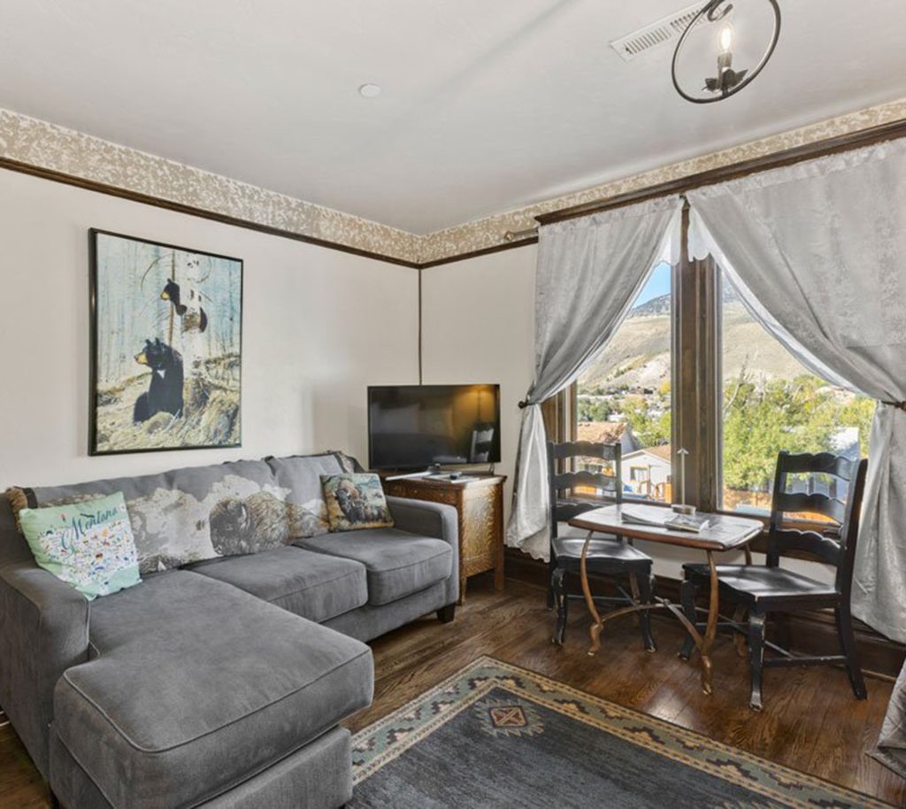 Cozy living room with a gray sectional sofa, animal-themed pillows, small TV, and framed bear artwork. Two black chairs and a table sit by a window with mountain views and light gray curtains.