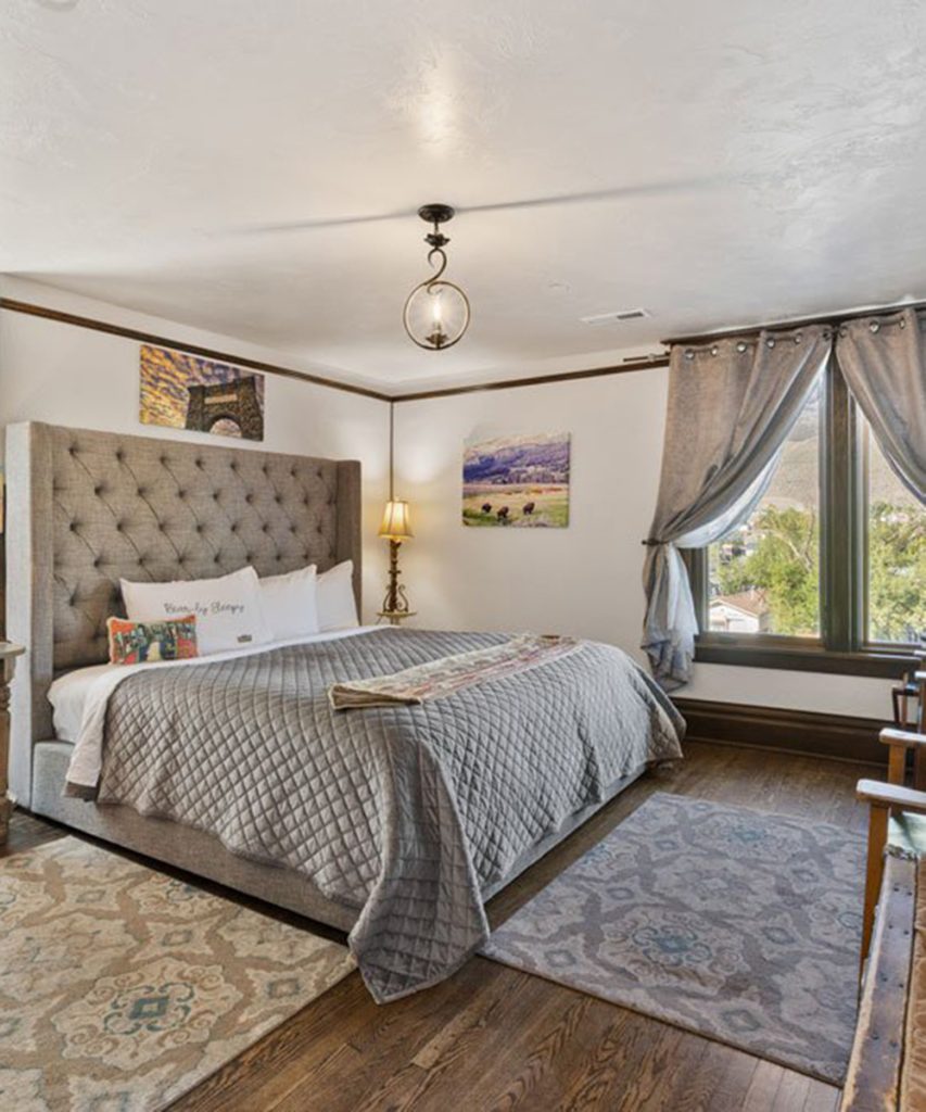 A cozy bedroom with a tufted gray headboard, gray quilted bedding, two rugs on wooden floors, artwork on the walls, a window with gray curtains, and a bedside lamp on a small table.
