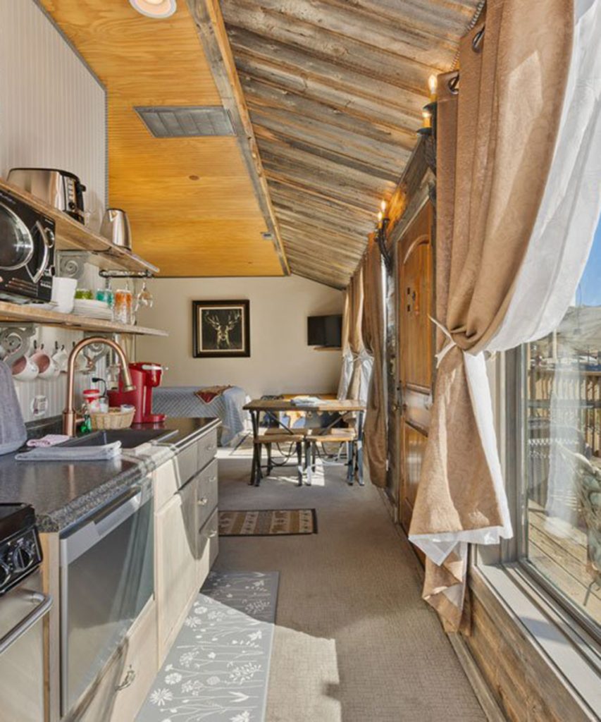 A cozy, narrow kitchen with rustic wooden ceiling, open shelves, modern appliances, and sunlight streaming through large curtained windows; a small dining area is visible at the end of the room.