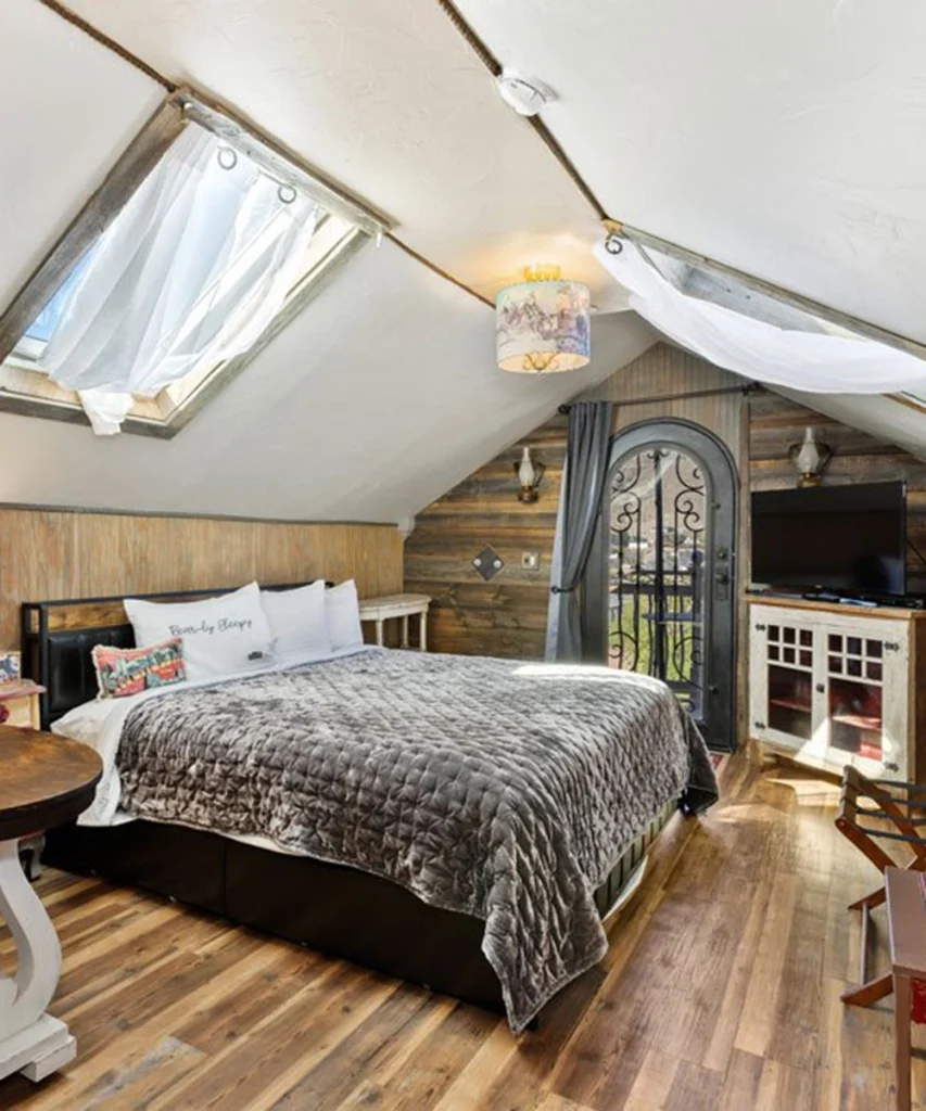 Cozy attic bedroom with sloped ceiling and skylight, featuring a large bed with gray quilt, rustic wood walls, wooden floor, TV on a cabinet, and a decorative arched door with wrought iron details.