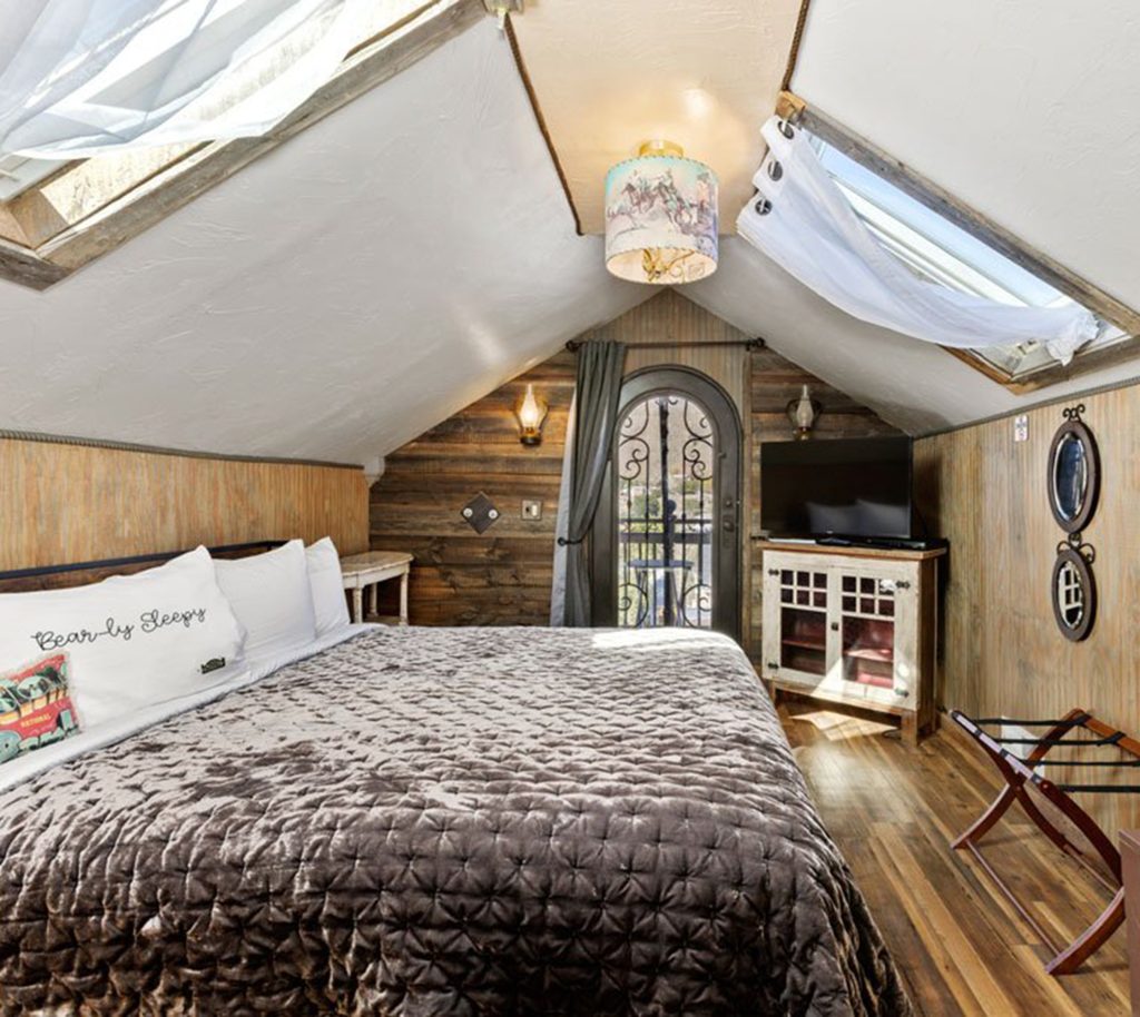 Cozy attic bedroom with a skylight, rustic wooden walls, a large bed with a gray quilt, decorative pillows, a TV on a white stand, and a wrought-iron door leading to a small balcony.