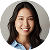 A woman with long black hair is smiling at the camera. She is wearing a light blue collared shirt and is pictured against a light gray background. The photo is cropped in a circular frame.