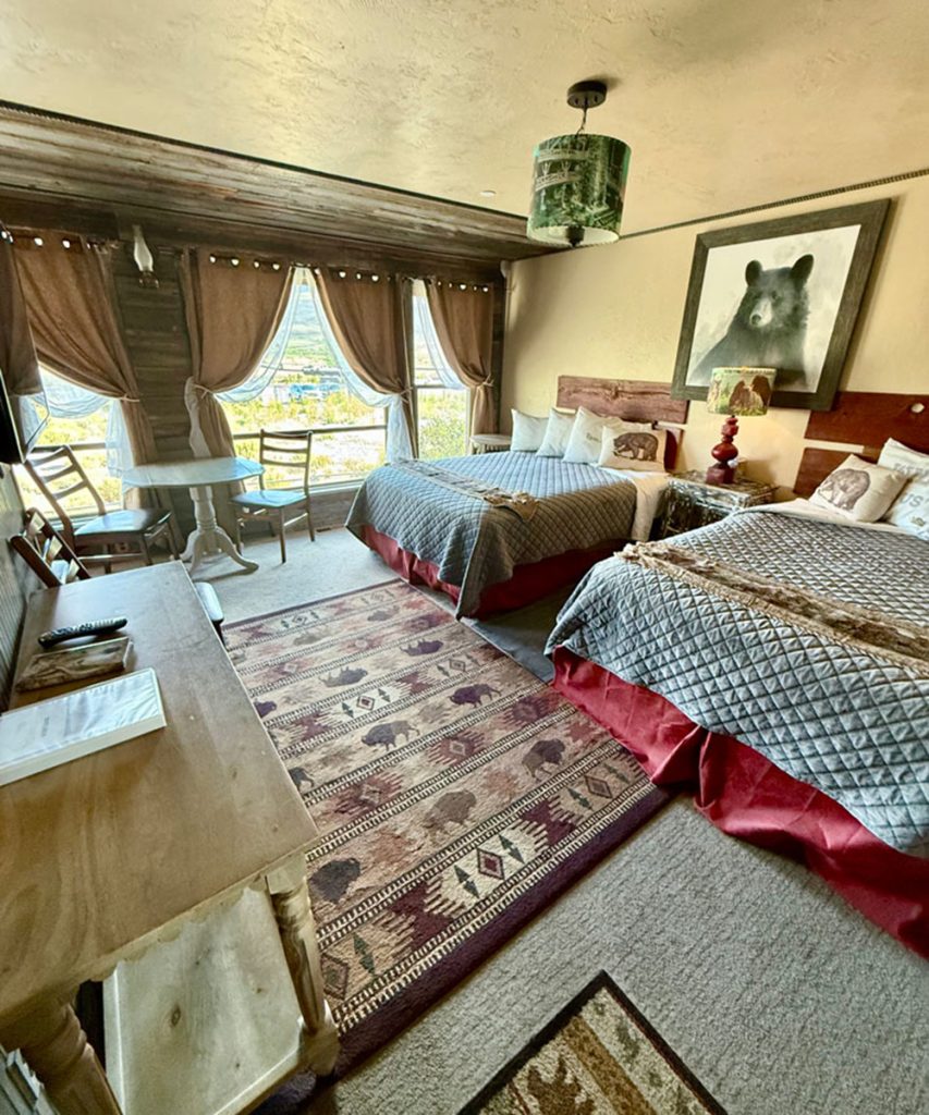 A cozy hotel room with two queen beds, rustic wooden furniture, a patterned rug, large windows with curtains, a small table and chairs, and bear-themed decor including a bear painting above the beds.