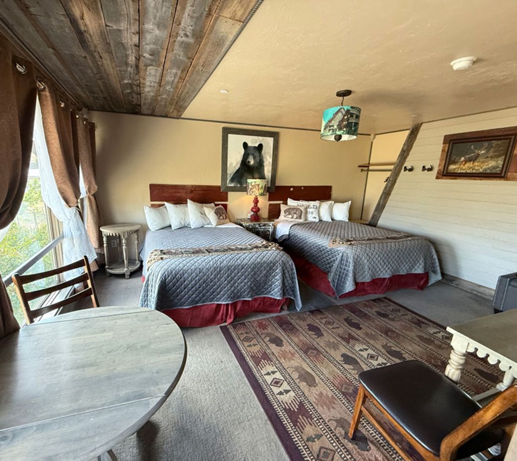 Cozy hotel room with two queen beds, gray bedding, a rustic wooden ceiling, a large bear painting above the beds, a round table with chairs, and natural light coming through curtained windows.