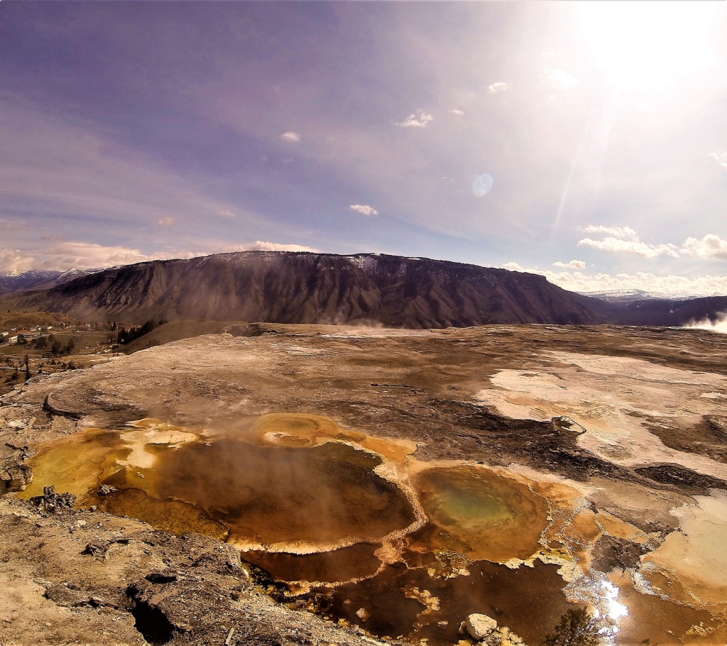 Steaming hot springs with colorful mineral deposits on rocky ground under a bright, sunny sky; distant mountains and wispy clouds are visible on the horizon.
