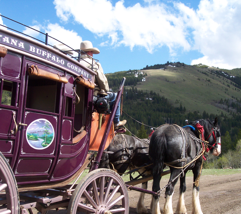 A person wearing a cowboy hat drives a maroon horse-drawn stagecoach labeled "Buffalo Company," with two black horses on a dirt road in a mountainous, forested landscape under a partly cloudy sky.