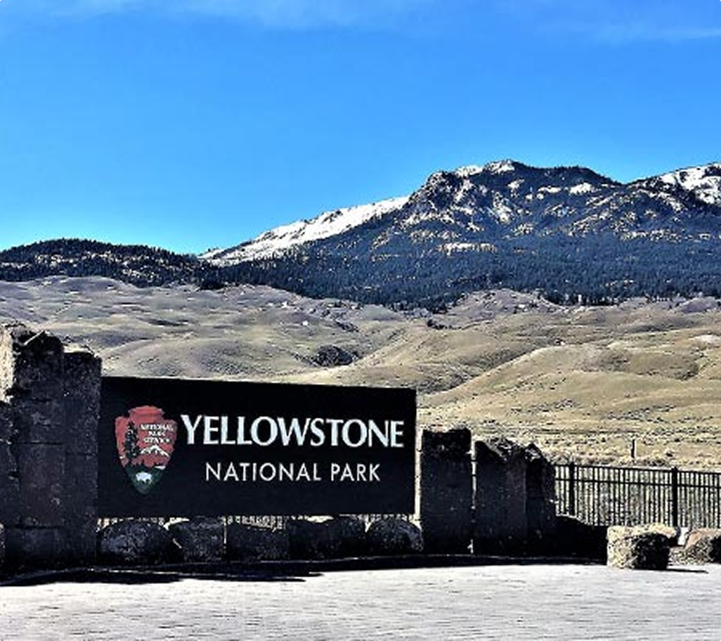 A large sign reading "Yellowstone National Park" stands in front of rocky hills and snow-capped mountains under a clear blue sky.