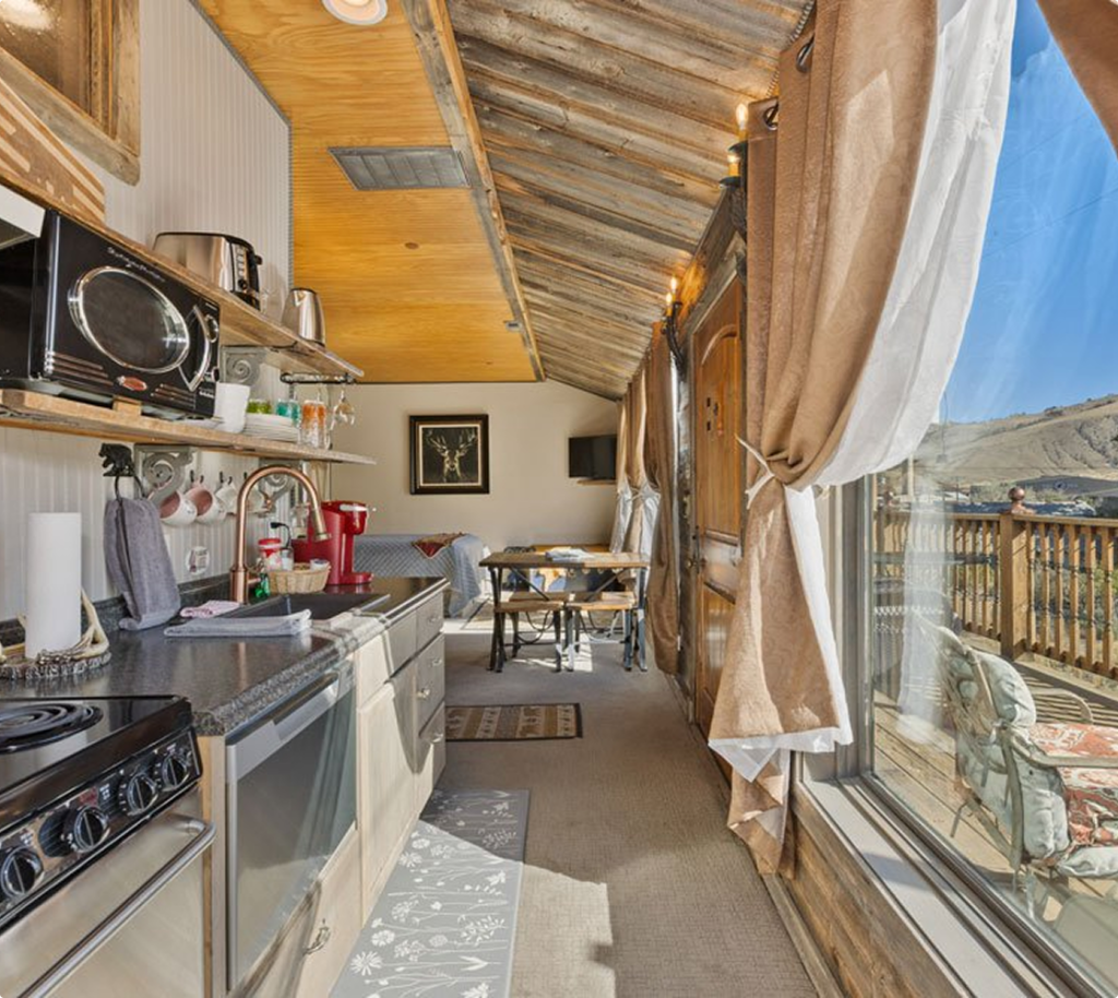 A cozy, narrow kitchen with rustic wooden ceiling, modern appliances, shelves, and a small dining table by large, sunlit windows with draped curtains, overlooking a scenic outdoor view.