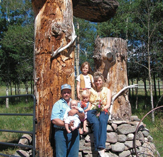 A family of four poses by large tree trunks outdoors; a man holds a baby, while a woman with a bouquet and a young girl sit on a rock wall. Green grass and trees are in the background.