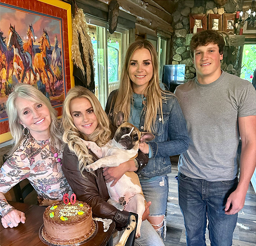 Four people stand inside a rustic wooden cabin, smiling by a table with a chocolate birthday cake. One woman is holding a small dog. A colorful painting of horses hangs on the wall in the background.