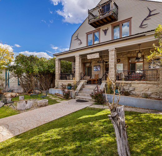 A rustic two-story house with a stone porch, balcony, and Western-themed decor sits under a blue sky. The yard has green grass, stone pathway, tree stumps, and a small tree in the foreground.