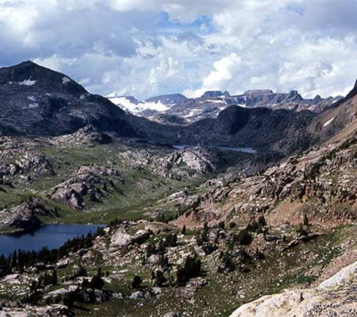 Mountain landscape with rocky terrain, green patches, scattered trees, and several small lakes. Snow-capped peaks rise in the background beneath a partly cloudy sky.